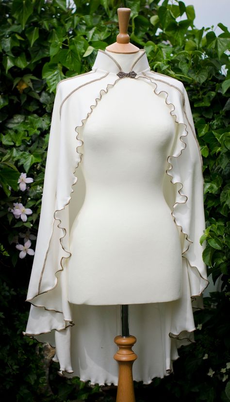 ~ White Driud Lily Capelet ~ Made to Measure ~ Original made in White material with Antique Gold thread & brass clasp ~  The Druid Lily Capelet is made from lovely stretchy cotton jersey with vertical striped detail, lettuce-edging & mandarin collar. It has a metal clasp closure at the throat, the clasps that I currently offer are a different style to those photographed on the cape. You can see the new style and colours in the last image. Perfect for Fanciful Faeries, Gothic Goddesses & Pixchiev