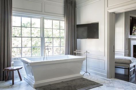 White and gray bathroom features walls adorned with decorative moldings framing windows dressed in gray curtains lined with a rectangular freestanding tub and a vintage style tub filler alongside a dark gray bath mat atop a marble herringbone tiled floor and a freestanding towel holder placed under a flat panel tv niche. Square Freestanding Bathtub, Rectangle Freestanding Bathtub, Rectangular Tub, Huge Bathtub, Master Tub, Bathroom Feature Wall, Georgiana Design, Bathroom Construction, Standing Bathtub