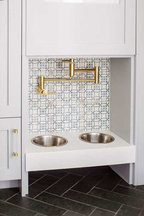 A brass swing arm pot filler is mounted to white and blue mosaic backsplash tiles over a built-in dog food bowl fixed above black herringbone floor tiles and between light gray-blue cabinets with glass and brass pulls. Black Herringbone Floor, Transitional Mudroom, Dog Station, Dog Food Station, Transitional Laundry Room, Mudroom Flooring, Grey Floor Tiles, Hydration Station, Bathroom Cabinetry