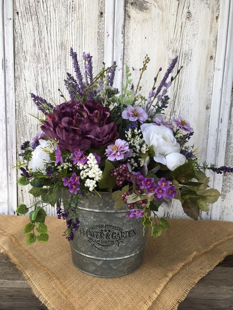 "A beautiful floral arrangement in pretty shades of purple, lavender and white with a variety of greenery constructed in a galvanized tin that reads \"Garden Supplies FLOWERS & Garden. 1967\". The centerpiece is perfect for spring, summer or year round. It can be viewed from all sides and displayed on your dining table, kitchen island, next to your fireplace or a very well protected area outdoors. Approximate Finished Dimensions: 18\" H X 15\" W tip to tip. Flower and Garden Container Dimens Purple And White Flower Arrangements, Planted Containers, Lavender Hydrangea, Purple Mason Jars, Purple Flower Arrangements, Purple Flowering Plants, White Floral Centerpieces, Country Christmas Trees, Fake Flower Arrangements