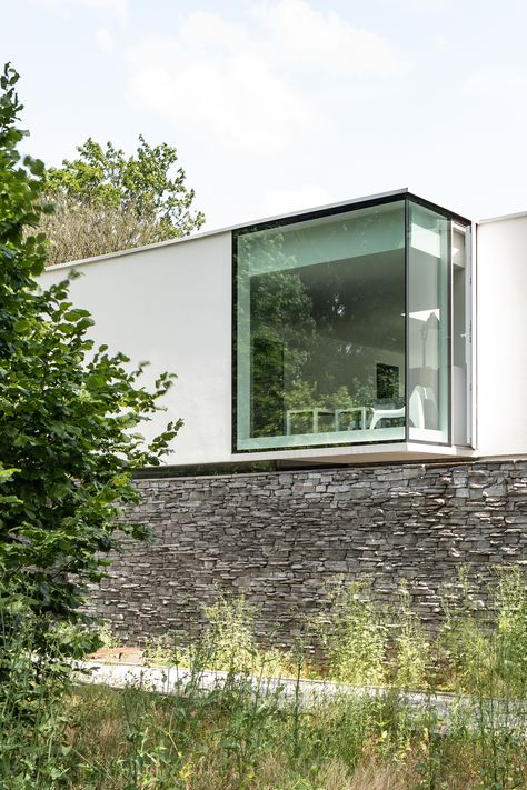 Located in a residential area on the outskirts of Brussels, this new house was designed to blend in discreetly while asserting its contemporary character. It is partially hidden behind the vegetation that has been preserved and is characterized by a very horizontal volumetry. A suspended volume ornates the front facade in which only a large bay window in the corner creates an interaction between the interior and the street. The large horizontal stone walls anchor the house to the land, ensure th Architecture Minimalist, Corner Window, Wooden Steps, Building Structure, Floor To Ceiling Windows, Ceiling Windows, Window Design, Home Construction, Architectural Elements