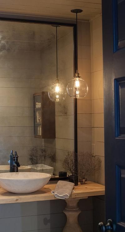 A black door opens to a gray powder room features walls clad in gray shiplap lined with a blond wood balustrade vanity topped with a marble bowl sink and bronze faucet under a black framed mirror lit by a single glass globe pendant. Powder Room Pendant Lights Over Vanity, Ensuite Pendant Lighting, Hanging Bathroom Pendant Lights, Hanging Light In Powder Room, Wall Lights In Bathroom, Oversized Mirror Powder Room, Pendant Light In Front Of Mirror, Powder Bathroom Pendant Lighting, Hanging Pendant Over Bathroom Vanity Mirror