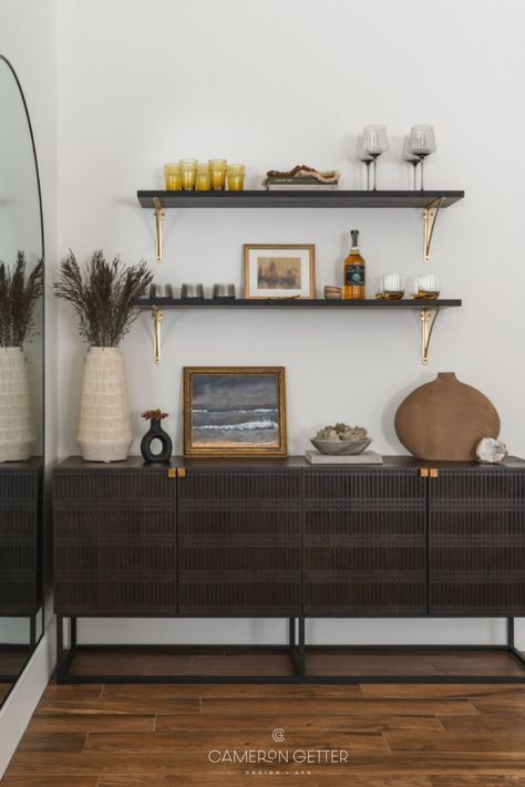 This home bar area refresh consisted of a carved mango wood sideboard, floating shelves with brass hardware, a black framed wall mirror, new glassware, artwork and styling accessories. Click the link on the image to view more from this project! #homebar #homebardesign #floatingshelves #wallmirror #framedartwork #glassware #stylingaccessories #vases #accessories Bar Cabinet With Floating Shelves Above, Credenza Bar Styling, Shelves Above Cabinets Living Room, Sideboard Bar Ideas, Buffet With Shelves Above, Bar Ideas For Home Living Rooms, Bar Cabinet Styling, Condo Entrance, Bar Credenza