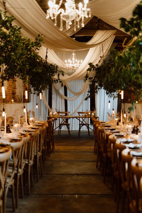 This is showing a barn wedding dining area. The overall vibes are rustic luxury. On the ceiling are glass chandeliers, white drapes hanging across the ceiling, directly above the wooden long dining tables are hanging boards with ivy on them. On the tables are white table runners, vases, candlesticks, tableware and glassware. In the middle of the two rows of dining tables is a sweet heart table at the end. Low Ceiling Draping Wedding, Wooden Crossback Chairs Wedding, Wedding Ceiling Drapery, Wedding Decor Hanging From Ceiling, Green Draping Wedding, Wedding Drapes Ceiling, Curtain Ceiling Design, Wedding Ceiling Decorations Draping, Black Draping Wedding