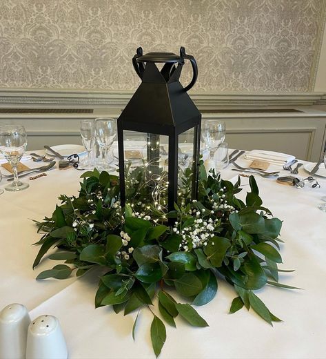 Wedding & Event Decor Hire on Instagram: “Simple but stunning wedding centrepieces. Black lanterns, fairy lights, foliage wreaths with a hint of gyp. #weddingflowers…” Lanterns With Greenery Centerpieces, Dark Green Greenery Wedding, Lantern Fairy Lights Centerpiece, Black Lantern Wedding Decor, Forest Green Centerpieces Wedding, Lantern Centerpieces With Fairy Lights, Black Lantern Table Decor, Wedding Table Decor Lanterns, Wedding Centerpiece With Lantern