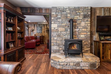 Stone accent, wood stove, and rustic hard wood flooring in a basement remodel by Judd Builders. Wood Stove Basement Ideas, Stone Hearths With Wood Stoves, Cabin Wood Burning Stove Ideas, Brick Wall Wood Stove, Pellet Stove With Stone Wall, Rock Wall Behind Woodstove, Wood Burning Stove Basement, Stone Hearth Wood Stove, Wood Stove In Basement