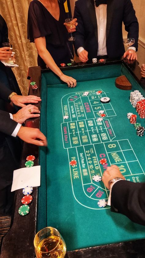 A craps table surrounded by tux covered hands, wine glasses, ladies in evening gowns. Wine glass in the lower corner, dice and chips on the table as play is in session Craps Table, Imaginative Writing, Casino Room, Surprise 60th, Green Room Decor, Casino Party Games, Casino Birthday, Casino Table, Poker Party