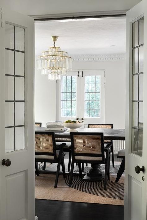 Dining space with leaded glass bi fold doors features cane dining chairs on a tan and black rug at a black oak dining table illuminated by an art deco chandelier. Chic Parisian Apartment, Bi Fold Doors, Living Spaces Inspired Home, Bamboo Dining Chairs, Cane Dining Chairs, Dining Room Console, Leaded Glass Door, Chicago Interior Design, Tulip Dining Table