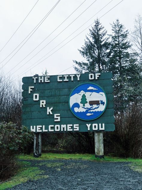 A sign reads "The city of Forks welcomes you". Trees surround the sign. Washington State Twilight, Twilight Forks Washington, Forks Washington Forest, Washington Forks Aesthetic, Forks Aesthetic Twilight, Fork Washington, Forks Washington Aesthetic, Forks Washington Twilight Aesthetic, Twilight Washington