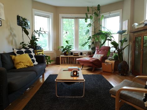 Vintage and retro living room with bay windows, crown molding, hardwood floors, black area rug, black couch, polka dot throw pillows, and a red leather lounge chair | Domu Chicago Apartments Couch Against Bay Window, Lounge Room Ideas Bay Window, Chair Couch Living Room, Apartment Bay Window Ideas, Bay Window Plants Living Room, Bay Window Apartment Living Rooms, Chicago Apartment Living Room, Leather Chaise Lounge Living Room, Bay Window Studio Apartment