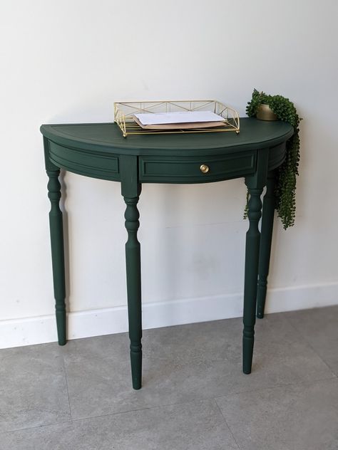 A rich dark,matte green semi circle table with long ornate legs contains a central drawer with a small gold knob on front. Table sits on white background and grey base and is furnished with a gold wire letter tray and small trailing plant which hangs off the right side of table top. Dark Green Table, Half Moon Table Decor, Green Tables, Painted Console Table, Console Table Makeover, Forest Green Console Table, Half Moon Table Makeover, Green Console Table, Painted Half Moon Table