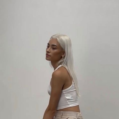 A photograph of a young Asian woman with tan skin and white hair, wearing a white cropped tank top. She faces three quarters left in front of a plain white background. Tan With Platinum Blonde Hair, Platinum Blonde Tan Skin, Platinum Hair On Tan Skin, Platinum Hair Olive Skin, Platinum Blonde Olive Skin, Platinum Blonde Hair On Tan Skin, Platinum Blonde Hair Tan Skin, Tan With Blonde Hair, Tan Skin White Hair