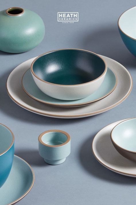 A medley of dinnerware and vase ceramics in off whites, blues, and teal on a dusty blue background. In the center there is a stacked Issaquah Dinnerware Set, a Sand glaze Dinner Plate under an Aqua Chocolate Salad Plate. The Cereal Bowl on top is Teal Sand. The Heath Ceramics logo in white is in the top middle of the image. Handmade Dinnerware, Heath Ceramics, Curated Home, Gadgets Kitchen Cooking, California Design, Dine In, Ceramic Dinnerware, Ceramics Pottery Art, Ceramic Houses