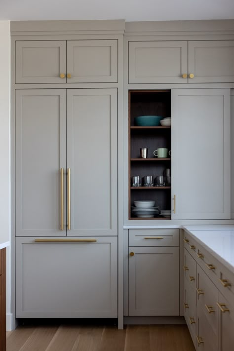 A streamlined kitchen corner boasts taupe Shaker style cabinetry with gold hardware, and chic white marble countertops. Cabinet doors slide to expose custom built-in shelves for easy organization, and the hidden refrigerator adds a sophisticated touch. Kitchen Shaker Cabinets, Hidden Appliances, Shaker Cabinets Kitchen, Gold Kitchen Hardware, Coastal Color Scheme, Yarmouth Maine, Open Shelving Kitchen, Gray Doors, Taupe Kitchen