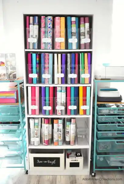 A white, tall bookshelf holds Cricut vinyl in clear storage bins organized by type. Labels make finding what you need easy and hassle-free. Vinyl Roll Storage Ideas Diy, Cricut Office, Craft Storage Ideas For Small Spaces, Office Craft Room Combo, Cricut Storage, Organizing Office, Craft Storage Solutions, Small Craft Rooms, Craft Spaces