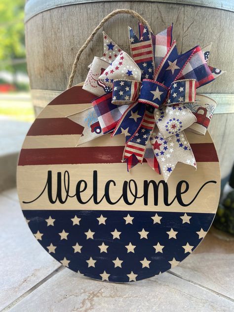 "Add a little patriotic farmhouse flair to your front door with this distressed Fourth of July or Memorial Day door hanger. Each door hanger is made from Baltic Birch wood and measures 16\" in diameter and .25\" deep and comes with a jute rope hanger. Back of sign is finished with stain or paint. Wording and designs are made from outdoor vinyl.  Greenery and bows are attached using staples.  Both front and back of signs are sealed for outdoor use, but it is recommended for indoor use or displayed on door under a covered porch or patio.   Each sign will vary slightly due to the way the wood will accept the paint and stain.  This is what will give each sign its unique and custom look.   Each sign is made at the time order is received.  Multiple quantities are listed so more than one person c Red White Blue Door Hanger, 4th Of July Door Hanger Diy, Patriotic Door Decorations, Fourth Of July Door Hangers, 4th Of July Door Hanger, 4th Of July Crafts, Fourth Of July Wreath, Patriotic Door Hanger, Americana Crafts