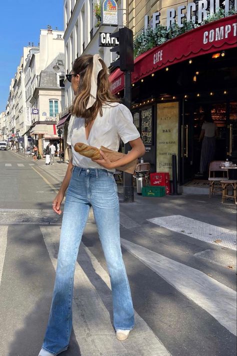 A girl is walking in the middle of the crosswalk in Paris, France. She is wearing light colored highwaisted skinny flared jeans. Her top is a wrap shirt that has a deep-V and it is fully white. Her hair is in a half-up ponytail hairstyle, and a white cream ribbon is tied to the top. Her hair is slightly wavy, and her head is looking to the side with a pair of black sunglasses. Her shoes are white and wicket brown. She is holding a baguette in her hand. The background is a corner coffee shop. Flared Jeans Summer Outfit, Bell Bottom Jeans Outfit Casual, Outfits With Bell Bottom Jeans, Bell Bottom Jeans Outfit Summer, Flare Jeans Outfit Summer, Light Denim Jeans Outfit, Flared Jeans Outfit Summer, Flare Jeans Outfit Spring, Conservative Clothing