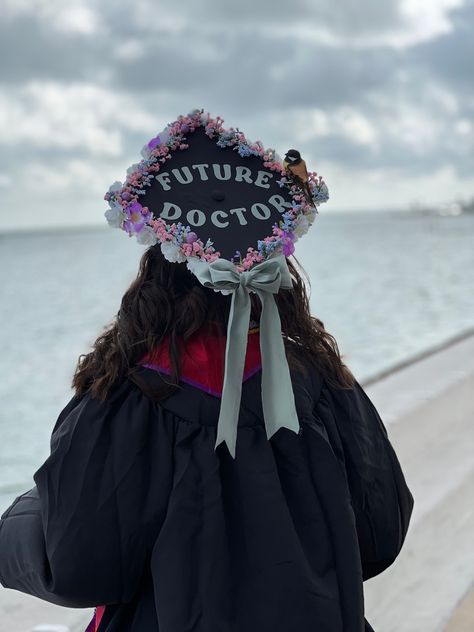 Picture of graduation cap with white roses on the bottom and pink, purple, and blue flowers on the top lining the cap, and surrounding the words “Future Doctor” in mint green in the center. A fake bird perches on the edge of the caps flowers. A large mint green bow in the same fabric and color shade as the words above is on the edge of the cap pointing towards her graduation gown Graduation Cap Designs Medical School, Future Doctor Grad Cap, Biology Graduation Cap Ideas, Future Doctor Graduation Cap, Graduation Cap Designs College Medical, Pre Med Graduation Cap, Medical Graduation Cap, Biology Graduation Cap, Graduation Cap Diy