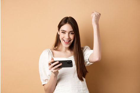 Girl winning on mobile phone. Happy woman raising hand up and scream yes with joy, achieve goal in smartphone app, standing on beige background. Girl With Mobile Phone In Hand, Phone Video, Happy Woman, Achieving Goals, Beige Background, Happy Women, Smart Phone, Scream, Mobile Phone