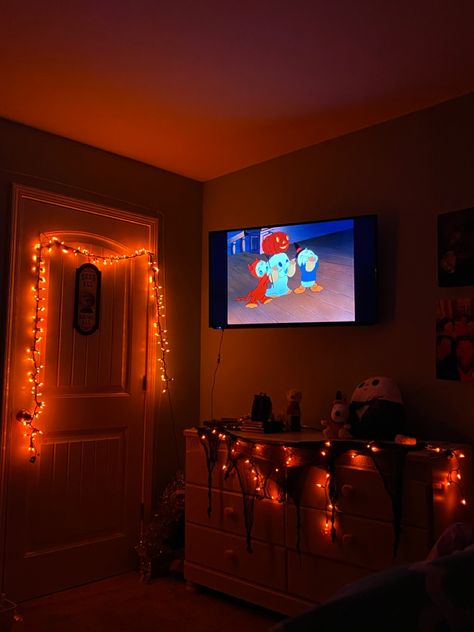 Orange lights decorating a room giving it a Halloween theme with jack the skeleton squishmallow and snoopy dressed as a pumpkin stuffed animal. Trick or treat mickey mouse special playing on tv. Halloween Lights Bedroom, Fall Themed Living Room, Halloween Theme Room, Halloween Room Ideas, Halloween Themed Room, Halloween Lights Indoor, Trick Or Treating Aesthetic, Cozy Halloween Bedroom, Fall Room Decorations