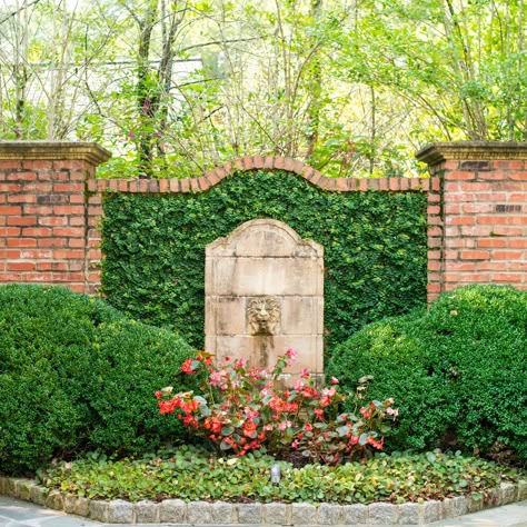 Here at Gardens to Love we take pride in making sure your garden is unique and special. This stone lion fountain among the sculpted bushes does a great job of illuminating the red brick behind it. Visit our website to learn all about the many features we could add to your space! #GardenstoLove #ATL Walled In Garden, Garden With Brick Wall, Brick Landscape Wall, Brick Walled Garden, Garden Features Focal Points, Garden Feature Wall, Brick Wall Garden, Garden Brick Wall, Brick Garden Wall