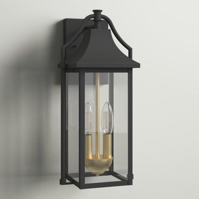 This 2-light outdoor wall sconce illuminates the way to your home with its soothing ambient glow and classic lantern frame made from wrought iron in a sleek black tone. The interior candelabra-style fixture comes in a gold finish and holds two dimmable bulbs（bulb not included）. Clear panels of beveled glass line all sides to complete this distinctively modern design. We love the dusk-to-dawn feature that automatically turns the light on when it gets dark and shuts off in the morning. Rated for w Front Porch Lanterns, Brass Outdoor Lighting, Porch Lanterns, Outdoor Wall Light Fixtures, Wrought Iron Decor, Porch Planters, Porch Swings, Black Wall Lamps, Outdoor Sconces