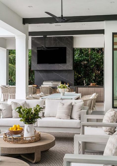 Covered patio features a light gray teak outdoor sofa with light gray teak lounge chairs at a round brown teak outdoor coffee table atop a tan rug and an outdoor kitchen area with gray rope counter stools at a marble topped island and a built in TV mounted over an outdoor cooktop. Outdoor Cooktop, Built In Tv, Outdoor Kitchen Area, Beach House Getaway, California Room, Tv Mounted, Dream Patio, Teak Lounge Chair, Sophisticated Bedroom