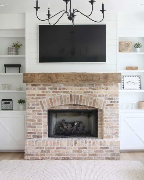 This white shiplap wall sports a coordinating white framed TV above a rough-hewn unfinished wood mantel and weathered brick fireplace. White built-in shelves and cabinets on either side display white, black, and wood decorations, while a modern black metal light fixture hangs from the ceiling in the center of the room. Fireplace Wall Ideas With Tv, Brick Fireplace White, Brick And Shiplap, Above Fireplace Ideas, White Stone Fireplaces, White Brick Fireplace, Faux Fireplace Mantels, Tv Over Fireplace, Brick By Brick