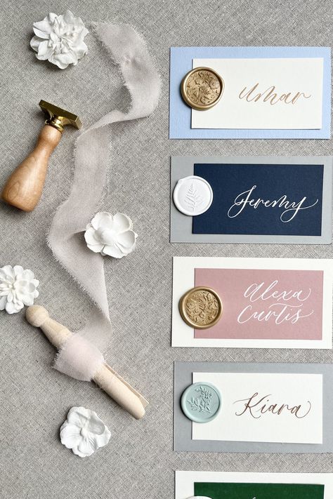 Different colored cardstock with hand-lettered calligraphy on a linen background with ribbon and flower accessories Wedding Name Cards With Wax Seal, Wedding Name Cards Diy, Wedding Guest Place Card Ideas, Wax Seal Place Cards, Creative Place Cards Wedding, Handwritten Place Cards, Gold Place Cards, Wax Seal Stamp Wedding, Calligraphy Place Cards