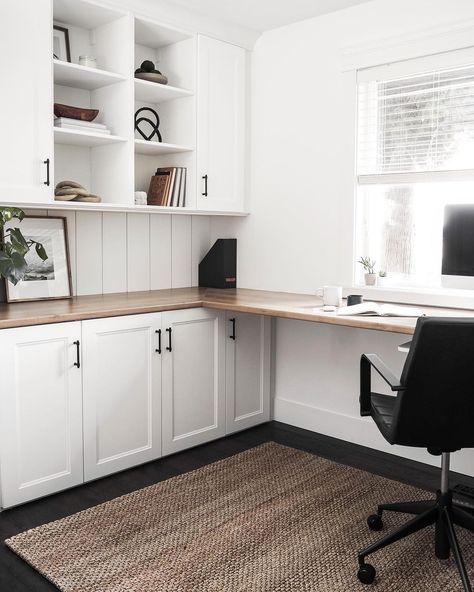 A spacious office with an L-shaped desk built into one wall features a black-and-white color scheme and a minimalist design. A woven rug covers the floor and a simple black office chair sits on top in front of a computer monitor and an open window with white blinds. Cubed shelves hold books, framed images, wooden baskets, and other modern decor. A plant, a larger print, and a small black magazine organizer adorn the desk. L Shaped Office Desk Ideas Layout, Built In L Shaped Desk, L Shaped Office, Built In Desk And Shelves, L Shaped Office Desk, Spacious Office, Basement Office, Office Built Ins, Minimalist Home Office