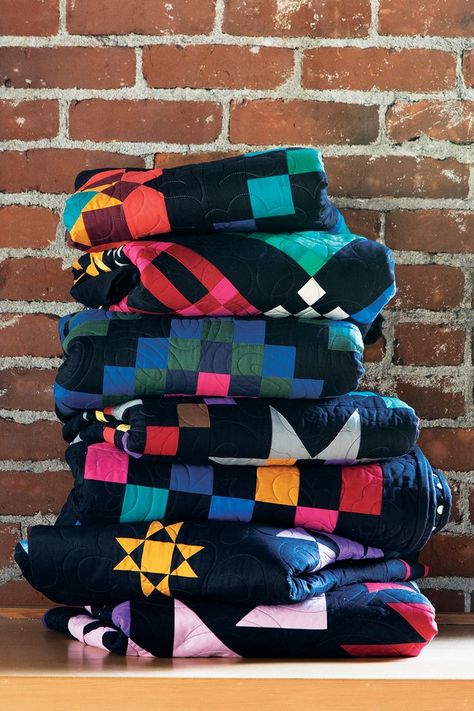 A stack of folded quilts are in front of a red brick wall. The quilts all feature black backgrounds and colorful patterns. Amish Quilt Patterns, Amish Quilt, History Of Quilting, Geometric Patchwork, Connecting Threads, Lemon Chiffon, Amish Quilts, Amish Recipes, Quilting Thread