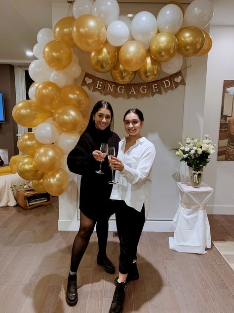 Two Indian women standing in front of decoration display made up or gold and white balloons and a banner that says engaged on it, cheersing with champagne glasses Casual Engagement Party Decorations, Small Engagement Party Ideas Simple, Gold Engagement Party Decor, Indoor Engagement Decorations, White And Gold Engagement Party, Simple Engagement Party Ideas, Elegant Engagement Party Ideas, Engagement Party Decorations Indoor, Classy Engagement Party