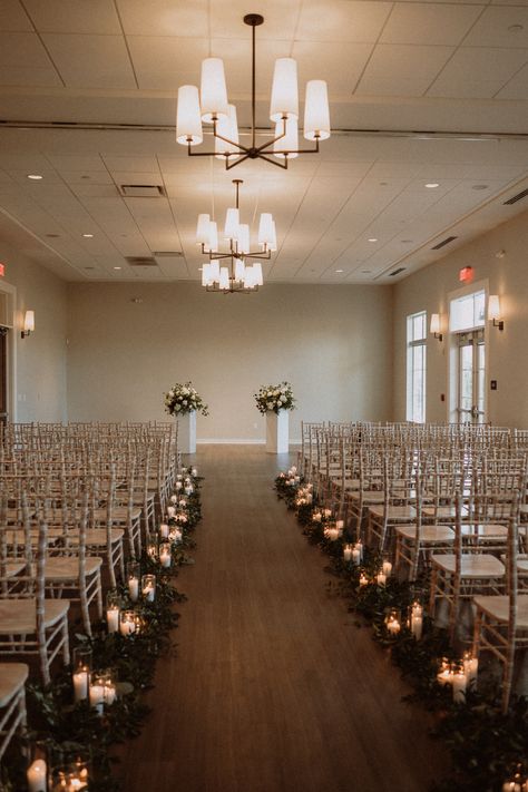 This photo shows an indoor wedding ceremony with greenery and clusters of pillar candles going down the length of the aisle. The ceremony chairs are white. The backdrop of the ceremony are two white columns with floral arrangements sitting on top. The fresh florals are white and have accents of greenery. Wedding Isle Candles, Candle Aisle Decor, Candle Aisle, Candle Isle, Wedding Alter Flowers, Wedding Aisle Candles, Tea Light Candles Wedding, Taper Candles Wedding, Aisle Candles