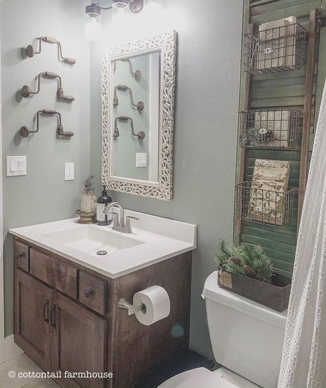 Three hand drills hang on the grey wall of this bathroom. A glass lamp fixture illuminates the ornate mirror beneath it. A small white countertop rests on a dark wood cabinet. Next to the cabinet sits a toilet with a small box of greens on it. Above the white toilet hangs three wicker baskets attached to a rustic green shelving unit. Distressed Bathroom Vanity, Gray Bathroom Walls, Chic Bathroom Decor, Bathroom Counter Decor, Gray Bathroom Decor, Farmhouse Vanity, Restroom Decor, Grey Bathroom, Bathroom Decor Apartment