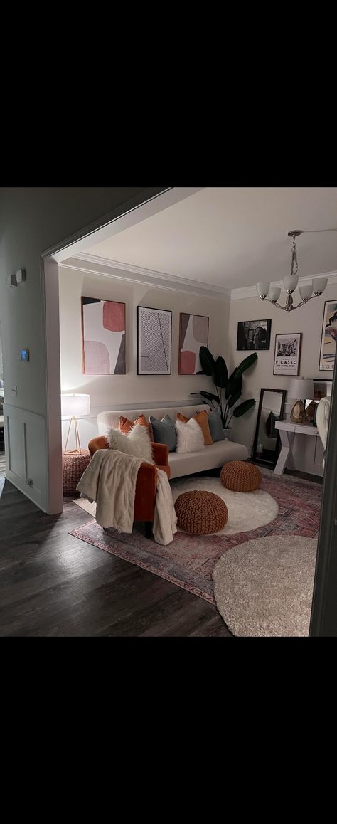 A room with dark brown hardwood floors, beige walls, and white ceiling. A pink rectangle rug covers most of the floor, with 2 small white circle rugs overlapping. On one wnd of the room, there is a white futon with white, orange, and Grey throw pillows; a fake tree on one side and a wicker table with lamp on top on the other. There is an orange side chair with white throw and pillow. Desk on opposite wall. On side wall, there is a mirror next to the fake tree and a side table with lamp and arts. Dining Room Space Alternatives, Closed Off Dining Room Ideas, Office Area In Dining Room, Small Dining And Sitting Room Combo, Dinning Room To Office, Formal Dining Room To Sitting Room, Apartment Den Ideas Small Spaces, Extra Dining Room Space Ideas, Turning Dining Room Into Sitting Room