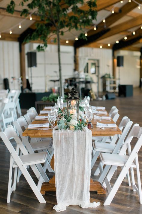 Wedding reception long wooden tables with white draped table runners, lanterns, and white red and greenery centerpieces with twinkle lights at wedding. #weddingreception #rusticweddingdecor #weddingdecor #weddingtablescape Wood Table White Chairs Wedding, Wood Tables With White Chairs Wedding, Lantern Centerpieces Long Table, White Table Runners Wedding, Wooden Table Decor Wedding, Wooden Tables Wedding, Wooden Table Wedding Decor, White Chairs Wedding, Lobola Decor