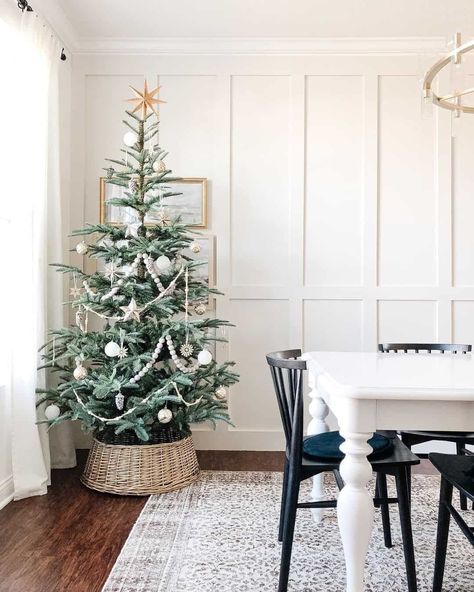 A Christmas tree with silver and white ornaments stands in a tree collar in front of a white board and battened dining room wall. A gold chandelier hangs over the white dining room table, while black chairs are placed on a patterned rug laying on the dark wood floor...   Image: caitlinmariedesign Noble Fir Christmas Tree, Neutral Christmas Tree, Minimalist Holiday Decor, Best Artificial Christmas Trees, Neutral Holiday Decor, First Christmas Together Ornament, Lamp Minimalist, Minimalist Christmas Decor, Neutral Christmas Decor