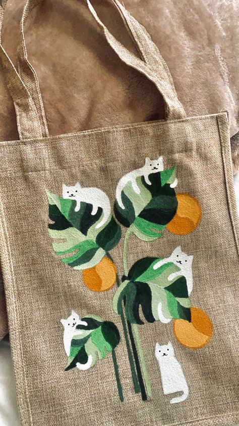 A beige tote bag has white cats hanging on leaves painted on it. Painting On Jute Bags, Paint Your Own Tote Bag, Canvas Tote Bag Painting Ideas, Toat Bag Painting Ideas, Painted Canvas Tote Bags, Bag Painting Design, Painting A Tote Bag, Hand Painted Canvas Bags, Drawing On Bags Ideas