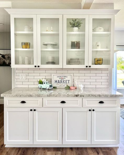 This neutral room features white kitchen cabinets with glass doors on the top section. A white subway tile backsplash contrasts with the gray marble countertop featuring rustic decor such as a wooden sign and spring themed ornaments. Glass Door Upper Kitchen Cabinets, Glass Front Kitchen Cabinets Ideas, Upper Kitchen Cabinets With Glass Doors, White Kitchen Cabinets With Glass Doors, Glass Kitchen Cabinets Decor, Glass Upper Kitchen Cabinets, Kitchen Cabinets With Glass Doors, Glass Cabinet Decor, Glass Door Hutch