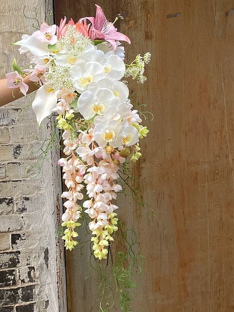 Indulge in the epitome of bridal luxury with this breathtaking faux bouquet. A cascading masterpiece which features a harmonious blend of exquisite white orchids, delicate pink lilies, and lush greenery, accented by the graceful allure of wisteria. The perfect complement to a modern, romantic, or boho-chic wedding, this bouquet exudes sophistication and timeless charm. The cascading design creates a dramatic and ethereal effect, while the high-quality faux florals ensure lasting beauty and elega Expensive Wedding Bouquet, Waterfall Bouquet Orchid, Lilly Flower Bouquet Brides, Wedding Bouquets Waterfall, Waterfall Orchid Bouquet, Lily Flower Bouquet Wedding, Orchid Waterfall Bouquet, Pink Lily Bouquet Wedding, Purple Orchid Wedding Bouquet