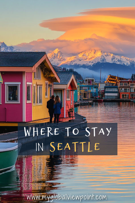 A couple walks along the waterfront by colorful houseboats in Seattle, with a stunning backdrop of snow-capped mountains and a dramatic sunset sky, highlighting one of the most beautiful places to stay in Seattle. Where To Stay In Seattle, Seattle Vacation, Seattle Neighborhoods, The Emerald City, Seattle City, Olympic National Park, Emerald City, Planning A Trip, Travel Itinerary