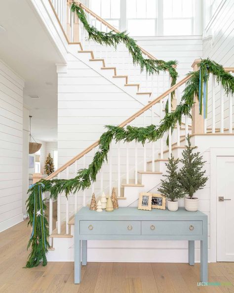 A coastal Christmas home entryway with white oak hardwood floors, white shiplap walls, a blue linen console table, garland on the staircase, and neutral and blue accents. Coastal Christmas Staircase, Coastal Christmas Wedding, Christmas In The Hamptons, Coastal Christmas Porch Decor, Christmas Coastal Decor, Coastal Holiday Decor, Lake House Christmas, White Shiplap Walls, Target Christmas Decor