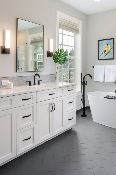 Hits of matte black plumbing fixtures, cabinet hardware and herringbone floor tile pop against the painted white cabinets and glossy quartz countertop and tub. The black and white color combination feels classic but still bold. Bonus! It’s the ideal backdrop for playing with color in artwork. Graphic Bathroom Floor Tile, Walk In Shower Black Fixtures, Hardwood Meets Tile, Primary Bathroom White Cabinets, Granite Backsplash Bathroom, White Vanity Grey Floor, Best Bathroom Floor Tile, Black Floors In Bathroom, Bathroom Ideas With White Tile