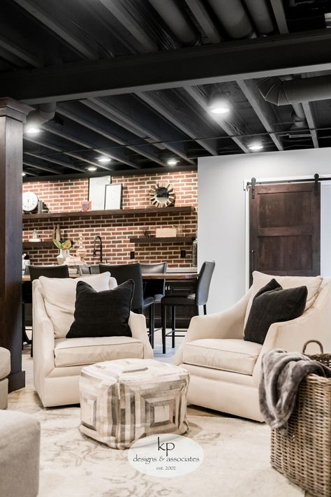 The client wanted to trade out their unfinished basement for a casual hangout where they could entertain and connect. The design inspiration was industrial with a touch of traditional to tie into the upstairs aesthetic. The entire lower level was renovated, adding a bedroom, bathroom, bar, and TV projector room. The finishes include traditional wainscotting and sconces in the TV area, an exposed ceiling, cold wine storage under the stairs... Basement Storage Closet Ideas, Upstairs Aesthetic, Black Basement Ceiling, Projector Room, Exposed Basement Ceiling, Concrete Basement Walls, Cheap Basement Remodel, Industrial Basement, Basement Movie Room