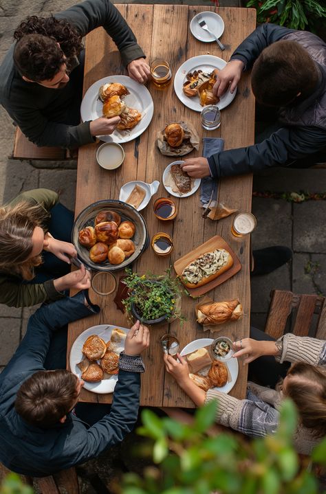 The image depicts a group of people gathered around a wooden table, engaged in what appears to be a communal and casual dining experience. The scene captures the individuals from an overhead perspective, focusing on their interaction with each other and the assortment of food laid out before them. There's a variety of dishes that suggest a brunch or breakfast setting, with items such as pastries, croissants, and possibly hot sandwiches, complemented by drinks that include coffee and glasses of Farm To Table Breakfast, Breakfast Family Table, Brunch With Friends Aesthetic, Overhead Perspective, Coffee Moodboard, Breakfast Setting, Gelatin Salad, Hot Sandwiches, First We Feast