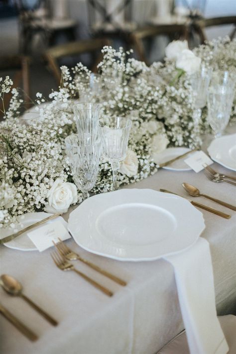 Image shows a closeup of a wedding table for guests. The linen is a neutral beige color with gold sileverware and a white plate setting. The centerpieces is a beautiful and simple floral arrangement that runs along the table of baby's breath and white roses. White Roses And Babies Breath Wedding, Babies Breath Wedding, Bridal Table Flowers, Engagement Party Table, Wedding Planning Boards, Bridal Party Tables, Table Arrangements Wedding, Minimalist Wedding Decor, Enchanting Wedding