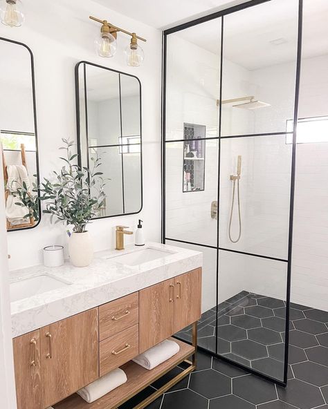 This bathroom boasts a large glass shower door, white walls, and a black hexagon tile floor. Brass lamps hang over black mirrors on the wall. A potted plant in a white pot sits on the white marbled countertop placed on the warm wood cabinets with brass drawer pulls. Black Hexagon Bathroom Floor, Black Hexagon Bathroom, Hexagon Bathroom Floor Tile, Black Hexagon Tile Bathroom, Hexagon Bathroom Floor, Hexagon Tile Bathroom Floor, Hexagon Bathroom, Wood Bathroom Cabinet, Hexagon Tile Bathroom