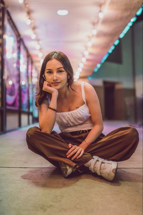 A night Photography photo of a beautiful girl with brown hair wearing a white tank top, brown pants, and white shoes, with glasses sitting down criss cross in front of bright lights Drawing Poses Sitting Criss Cross, Women Sitting Cross Legged, Pose Reference Criss Cross, Character Sitting Criss Cross, Sitting Poses Criss Cross, Sitting Cross Legged On Floor, Sitting Pose Reference Criss Cross, Cross Cross Applesauce Pose, Person Sitting Photography