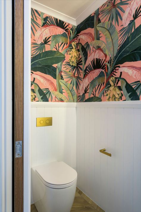 Fun Maximalism Powder Room | Maximalists and traditionalist unite in this little slice of the tropics. Herringbone laid wood-look tiles and VJ panelling speak of timeless style, while the pink and green banana palm wallpaper bursts with colour and unique expression. There’s so much to love in this separate toilet; in-wall cistern with plated buttons, brushed gold tap ware, cement Nood Co. hand basin and a stage stealing wall sconce. Toilet With Panelling And Wallpaper, Bathroom Ideas Quirky, Colourful Bathroom Wallpaper, Small Toilet With Wallpaper, Colourful Small Toilet Room, Pink And Green Downstairs Toilet, Tropical Wallpaper Powder Room, Colorful Toilet Design, Bold Toilet Room