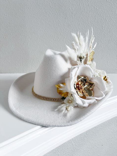 An elegant light Ivory felt hat embellished with decorative accents. Adorning the side of the hat with two large  fabric rose and gold brass flowers, accompanied by a variety of elements that add a touch of glamour. A gold rhinestone trim chain lies gracefully around the brim, giving the hat a luxurious feel.  Details -Light Ivory Fedora Cowboy hat -Off white/light Ivory florals in silk fabric  -Variety of Dried Flowers  -Gold brass florals  -Adjustable hat  One size fits most. Bridal Rancher Hat, Fancy Cowboy Hat, Western Bride Hat, Cow Boy Hats, Hat Decorating Ideas, Embellished Cowboy Hat, Western Cowgirl Outfits, Cowgirl Hats Western, Western Bride